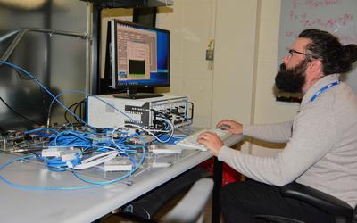 Man with beard and glasses in light grey sweater sitting on the right looking at computer screen on the left, with mass of blue wires in the foreground.