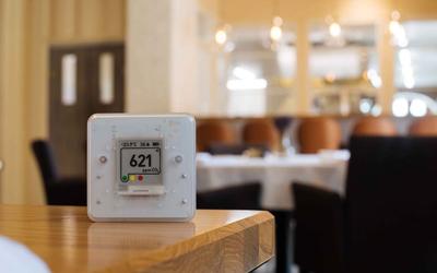 A white, square plastic CO2 meter sits on a table in a restaurant setting, reading 621 ppm.