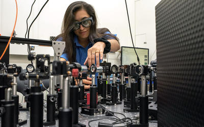 Woman with long dark hair wearing safety goggle stands in front of a table that has a series of lenses on stands