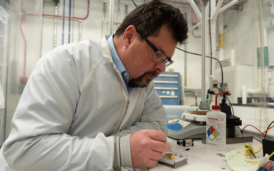 John Kitching leans over a table in the lab, using tweezers to point to part of a chip-scale device in a clear plastic box. 