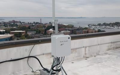 On a rooftop overlooking a waterfront town, an equipment box is attached to a pole anchored with concrete blocks. 