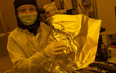 NIST researcher Jessie Zhang, wearing a protective suit, mask, and safety glasses holds up a piece of foil in the lab. 