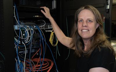 Jeanne Quimby crouches next to a rack of computer equipment with many different colored cord connections. 