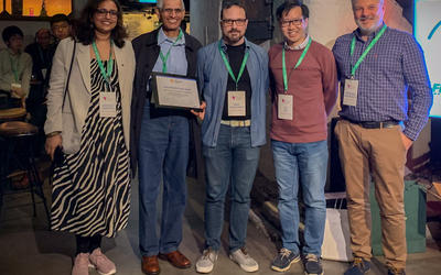 Five people dressed in business casual and wearing conference badges on lanyards pose with a framed award certificate.