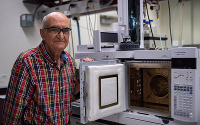 Miral Dizdaroglu wears safety glasses as he poses in the lab next to an open spectrometer, which looks a little like a microwave with extra buttons. 