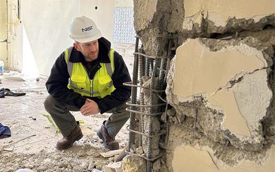 A man in safety gear crouches to examine a damaged wall inside a building after an earthquake. 