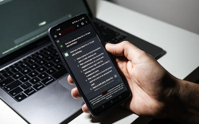 In front of a laptop computer, a hand holds a cell phone that has a conversation with generative AI on the phone screen.