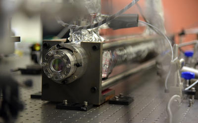 A scientific device attached to a lab table has a round, porthole-like opening on one end of a cylinder wrapped in foil. 