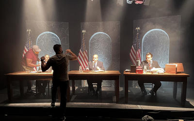 A man faces away from the camera, gesturing as he speaks to three actors seated behind desks on a film set. 