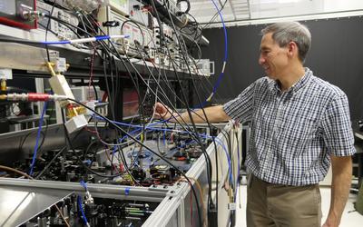 Tasshi Dennis stands in his lab and reaches toward a countertop full of equipment with lots of wires and circuitry.