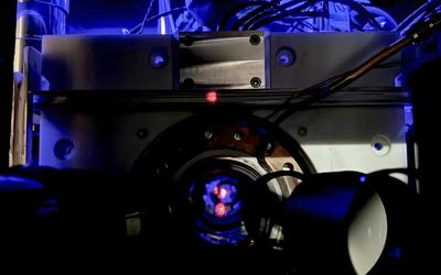 Close-up of a scientific instrument with wires and a circular metallic component, illuminated by blue light, featuring a small red dot at the center.
