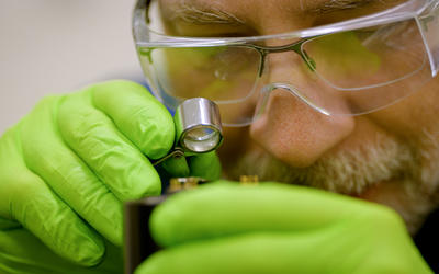 A researcher wearing green gloves and safety glasses over his regular glasses holds a small magnifying glass close to his face to look at a hard drive.
