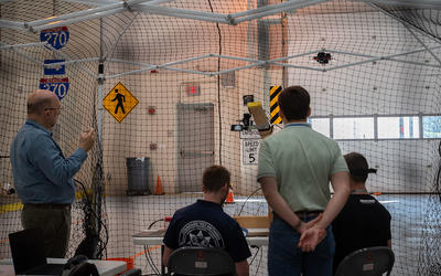 Four men are protected by a net structure as they look away from the camera toward a drone flying in a large high-ceilinged room with street signs on the walls.