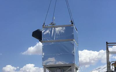A rectangular box coated with reflective foil hangs from cables over a parking lot.