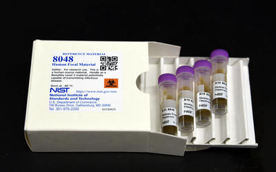A white box labeled "8048 Human Fecal Material" lies open, with a tray of four plastic vials pulled out. 