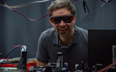 Krister Shalm, wearing safety glasses, smiles over a lab table covered with electronics equipment.