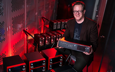 Scott Davis poses smiling and holding a frequency comb console. Other similar electronic products are stacked on carts around him. 