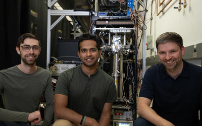 Three male researchers pose smiling in front of a large, complex scientific device in a lab.