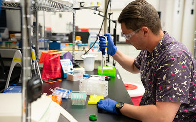 Researcher in safety glasses and gloves sits at a lab bench working with small test tubes.