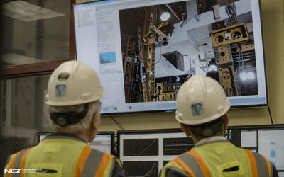 Two people in hard hats, shown from behind, look at a large screen showing concrete slabs and testing equipment.
