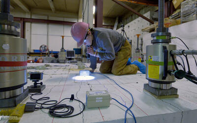 A researcher in a hard hat kneels on a concrete slab and uses a flashlight to examine it closely.