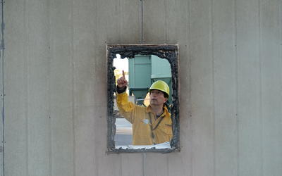 A man wearing a yellow hardhat and fire-resistant jacket points to burnt frame of a window.