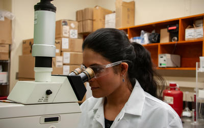 A woman wearing a lab coat and safety glasses stares into a large microscope.