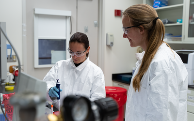 A researcher in a lab coat and safety glasses uses lab equipment as another researcher looks on. 