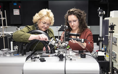 Angie Hight Walker and Amber McCreary stand side by side in the lab adjusting a complex scientific device.