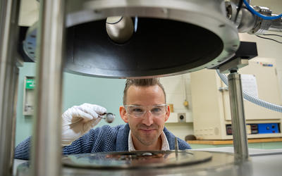 Andrew Iams wears safety glasses as his face is framed in an opening between two horizontal pieces of equipment in the lab. 