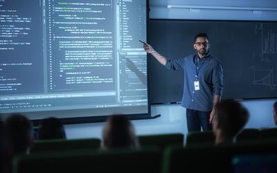 A young male teacher stands at the front of a darkened classroom, pointing at computer code on a large screen. 