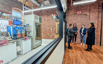 Four people stand talking in a converted factory space near a large interior window through which a complex, high-tech manufacturing line is visible. 