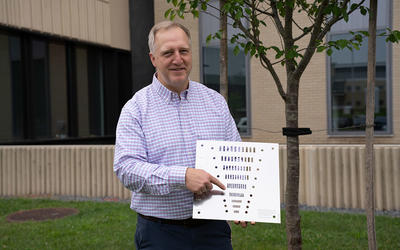 Jack Glover poses outside on the NIST campus, holding a flat square object with holes of varying sizes. 