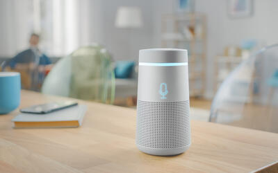 A cylindrical smart speaker sits on a wooden table in a living room. 