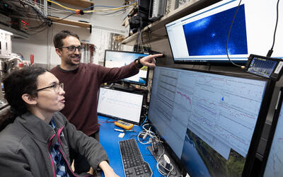 A woman with glasses and a long ponytail is seated at a computer with four display screens. A man with glasses stands beside her and points at the uppermost display screen. In the middle of that screen is a small blue glowing dot. The lower screens display data from the experiment.