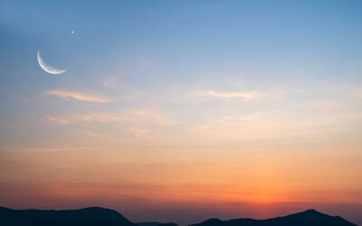 A sliver of moon is visible in the sky during sunset over mountains.