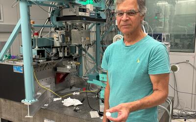 Jim Cline stands in front of a large scientific device in a lab, holding a small white disk.