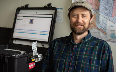 Gary Howarth poses for a photo in his office, wearing a NIST baseball cap, with a ruggedized laptop on the counter next to him and a map on the wall behind him.. 