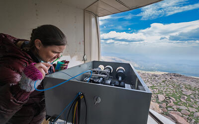 A researcher peers into a box of scientific equipment near an opening in the wall showing the view down the mountain. 