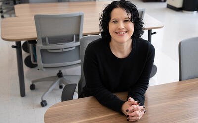 Kelly Sauerwein poses sitting at a table in the NIST library, leaning forward and smiling. 