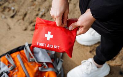 A person holds a red pouch labeled First Aid Kit as they stand over a travel bag on sandy ground.