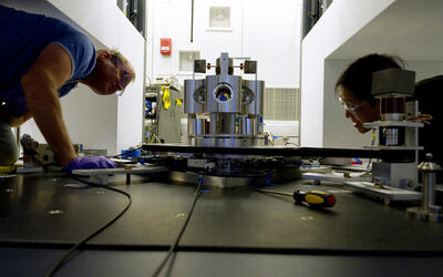 Wide shot shows two researchers on either side of a scientific device made of metal cylinders, peering closely at it. 
