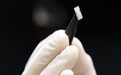 Close-up of a gloved hand holding tweezers that are grasping a tiny rectangular chip. 