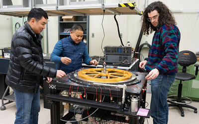 Three warmly dressed researchers stand around a scientific device with a large circular piece lying flat on top. 