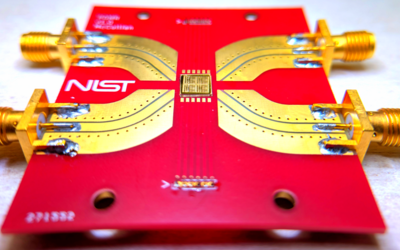 A bright-red rectangle with a NIST logo holds a gold-colored chip in the center with metal paths leading to connectors at the edge of the rectangle. 