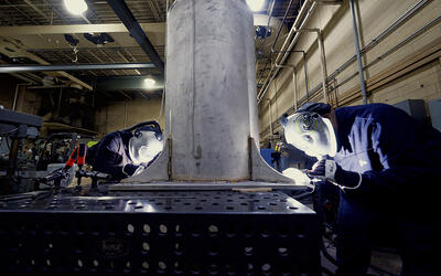 Two people wearing welding masks and other safety equipment bend over a table to work on a large metal cylinder.