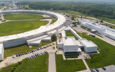 An arial view shows part of the huge enclosed loop of an accelerator, with attached buildings and parking lots.