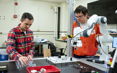 Two young men stand a lab table fitted with a white articulated robot arm.