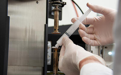 Researcher holds a small white tube filled with wet ceramics and points it toward lab equipment.