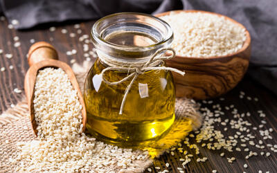 Bottle of sesame oil on a wooden table.  Sesame seeds are in a bowl and spoon next to the bottle and also sprinkled on the table.